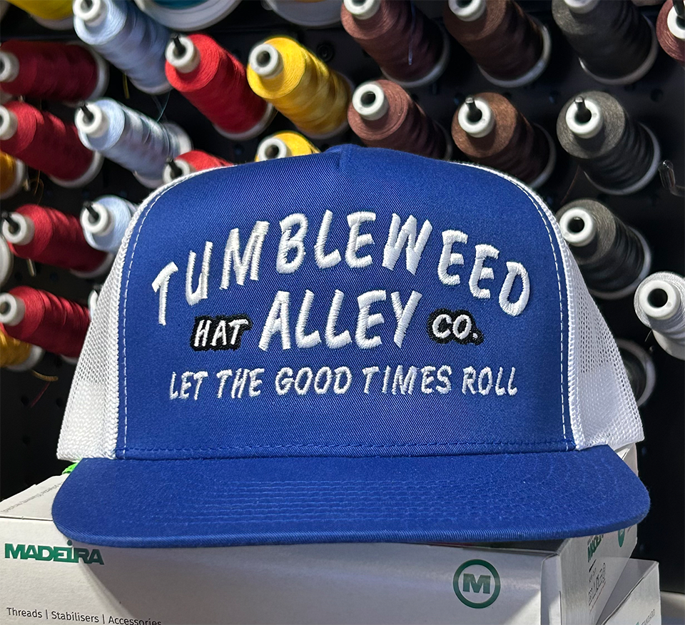 Blue cap with 'Tumbleweed Alley' text against a backdrop of spools of thread